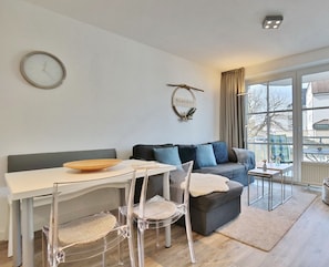 Una sala de estar moderna con comedor, un sofá gris con cojines azules, una mesa de madera y una silla de vidrio transparente.