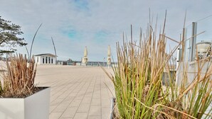 A paved walkway with planters containing tall grasses, a distant view of a building, and a clear sky.