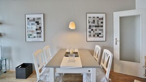 A dining room with a rectangular table, white chairs, a pendant light, and two framed abstract artworks on the wall.