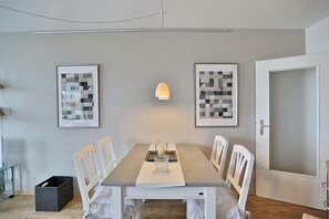 Une salle à manger avec une table rectangulaire, des chaises blanches, un luminaire suspendu et deux œuvres d’art abstraites encadrées accrochées au mur.