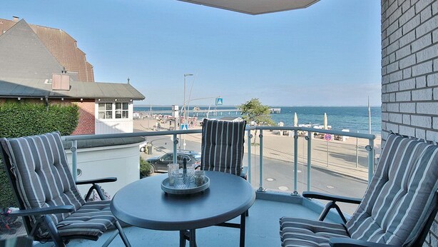 A balcony with two striped chairs and a small round table, overlooking a beach and the sea.