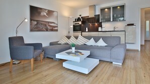 A modern living room with a grey sofa, a white coffee table, and a grey armchair. A painting is on the wall, and a kitchen is visible in the background.