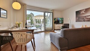 A modern living room with a grey sofa, a dining table with chairs, a TV on the wall, and a large window with a view of the outdoors.