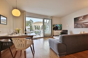 A modern living room with a grey sofa, a dining table with chairs, a TV on the wall, and a large window with a view of the outdoors. - Domizil am Meer Apartment 13 - Domizil am Meer (Timmendorfer Strand)