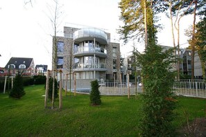 Un immeuble résidentiel moderne à plusieurs étages, doté d’un balcon incurvé, entouré d’une pelouse bien entretenue et d’arbres.