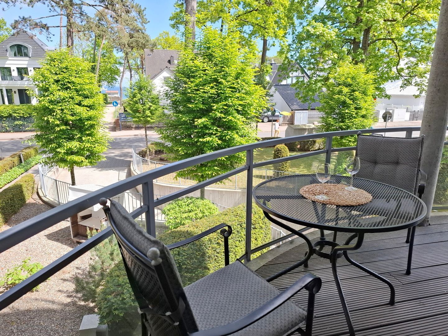 A balcony with a round table and two chairs, overlooking a street with trees and houses.