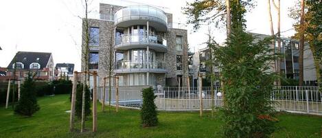 A modern multi-story residential building with a curved balcony, surrounded by a well-maintained lawn and trees.