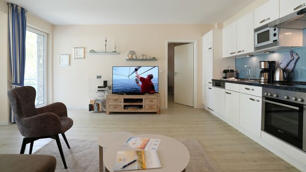 A modern living room with a flat-screen TV, a comfortable armchair, a small coffee table with magazines, and a kitchen area with a microwave and oven. - Home by the Sea Apartment 9 - Home by the Sea (Timmendorfer Strand)