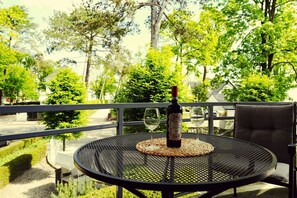 Ein runder Metalltisch mit einer Weinflasche und zwei Gläsern auf einer gewebten Tischunterlage, auf einem Balkon mit Blick auf Bäume und eine Straße.