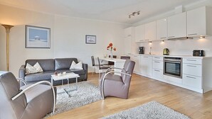 A modern living room with a grey sofa, a coffee table, and a dining area with chairs. A kitchen with white cabinets and appliances is visible in the background.
