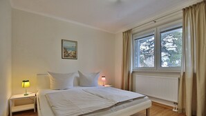 A bedroom with a bed, two bedside tables, a night lamp, a window with curtains, and a painting on the wall.