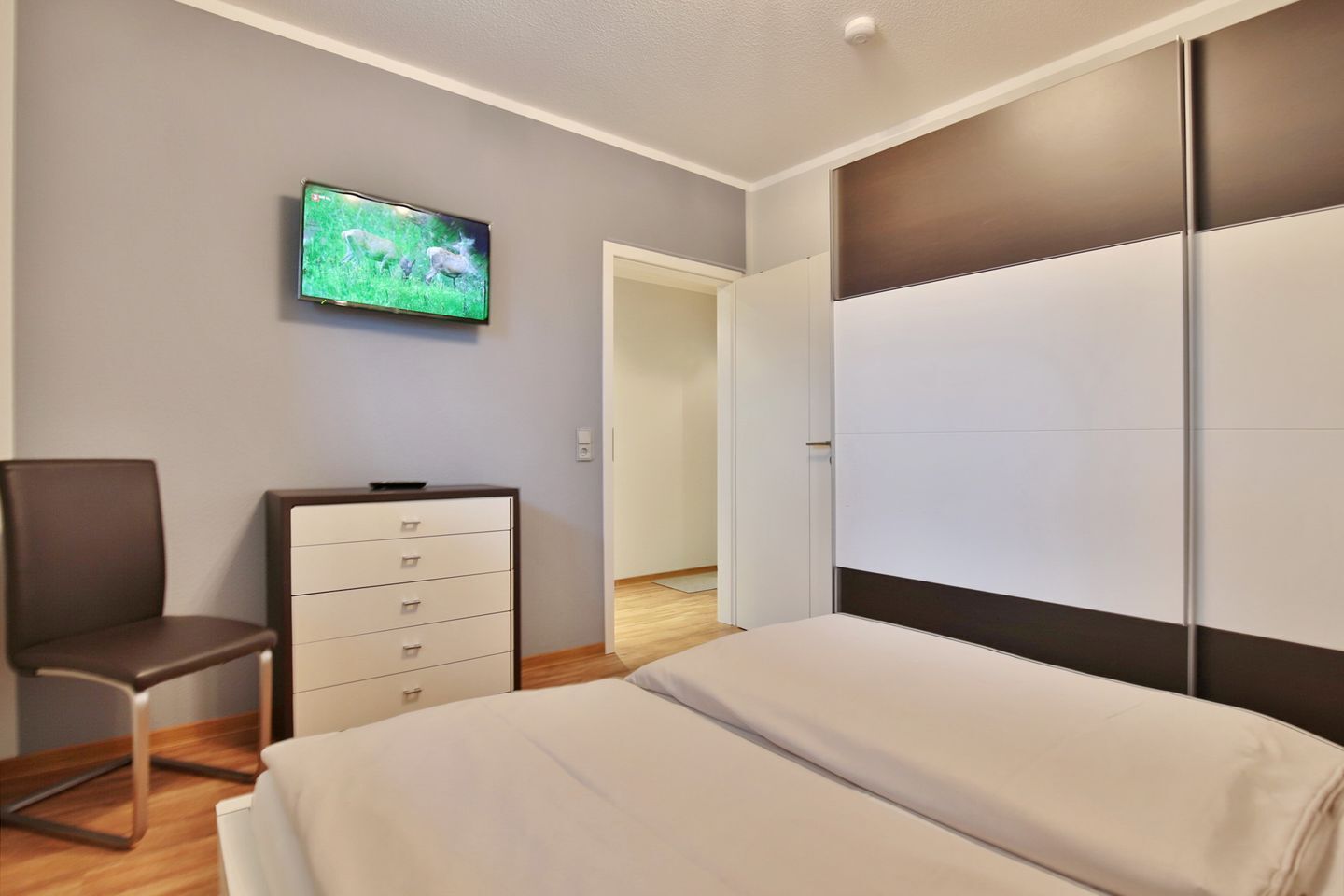 A bedroom with a bed, a dresser, a chair, and a TV on the wall.
