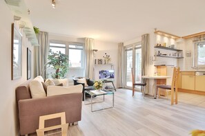 A modern living room with a sofa, coffee table, and TV. There is a dining area with a table and chairs. The room has large windows and a kitchen area with cabinets and a countertop.