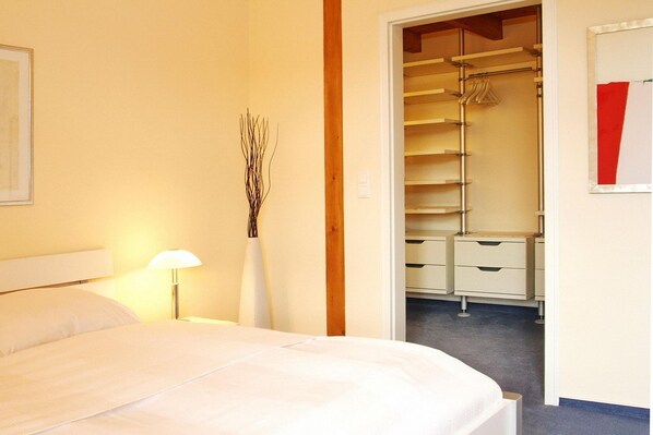 Habitación de hotel con cama, lámpara de noche, un florero con ramas y armario con ropa colgada.