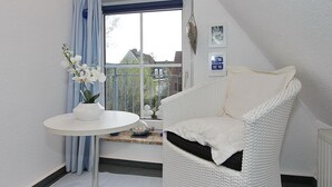 A corner room with a white wicker chair, a small round table with a white vase, and a window with a view of houses.