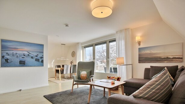 Una sala de estar moderna con un sofá gris, una mesa de centro de madera y un sillón gris. Hay una zona de comedor con una mesa y sillas, un ventanal con vistas, y obras de arte en las paredes.