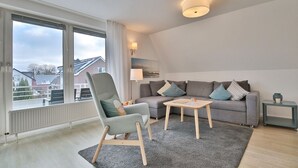 Una sala de estar moderna con un sofá gris, una mesa de centro de madera y un sillón gris. Se ve una vista de casas a través de amplios ventanales.