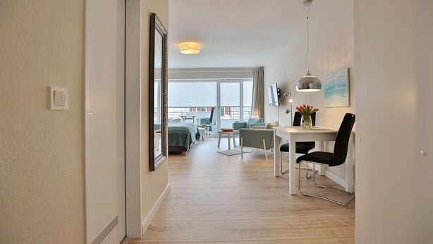 A modern apartment with a wooden floor, a dining area with a table and chairs, a sofa, and a large window offering a view.