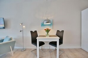 Et moderne spiseområde med et hvidt bord, to sorte stole, en vase med blomster, en hængende lampe og et maleri af en strandscene.