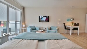 A modern hotel room with a large bed, a seating area with a table and chairs, a TV mounted on the wall, and large windows.