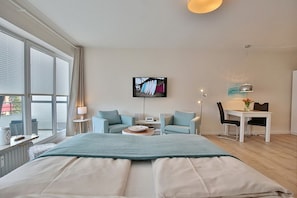 A modern hotel room with a large bed, a seating area with a table and chairs, a TV mounted on the wall, and large windows.