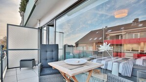 A modern balcony with wicker furniture, a glass table, and a view of neighboring buildings.