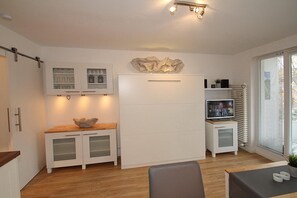 Une cuisine moderne avec des meubles blancs, un plan de travail en bois et un four encastré. On y trouve un petit coin salon avec une table et des chaises, ainsi qu’un téléviseur fixé au mur.