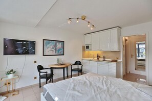 A modern bedroom with a bed, a desk with chairs, a TV, and a kitchenette.