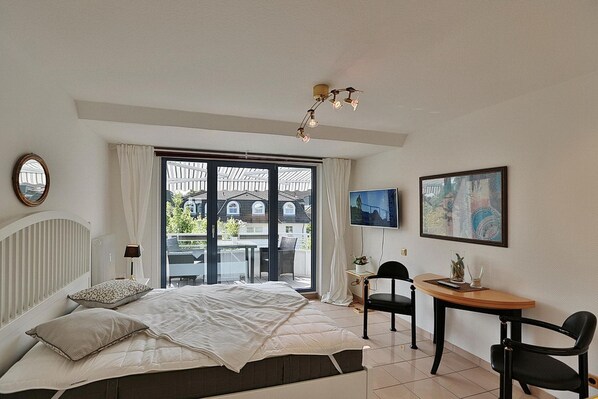 A bedroom with a bed, a desk, a chair, a TV, and a balcony with a view of houses.