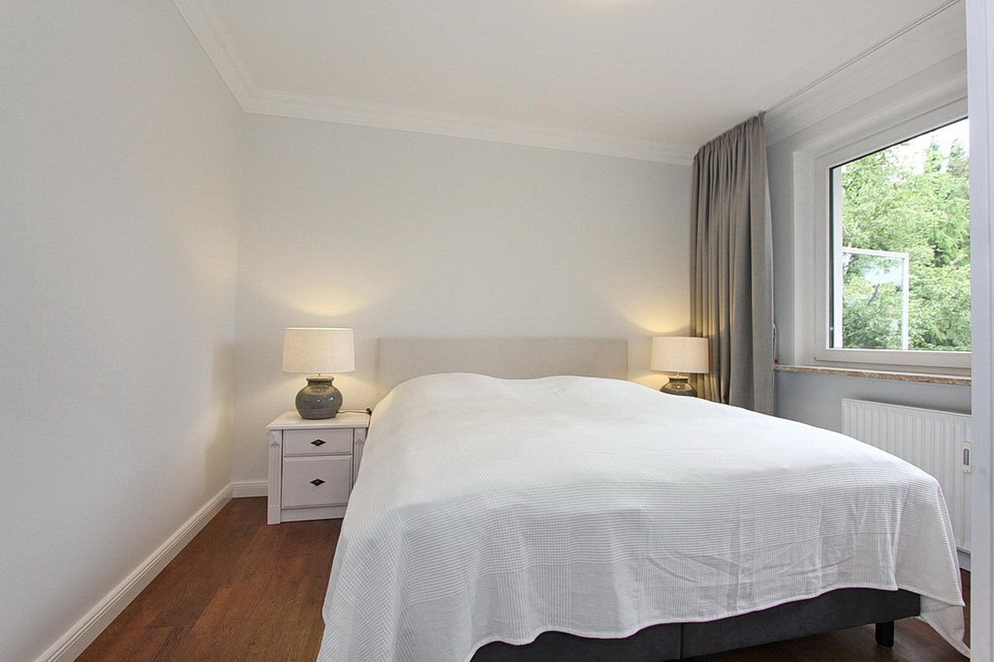 Una camera da letto con un letto grande, due comodini con lampade, una finestra con tende e un termosifone.