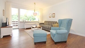 Un salon comprenant un canapé, un fauteuil, un pouf et une table basse. On y trouve également un radiateur, un téléviseur et une fenêtre avec des rideaux.