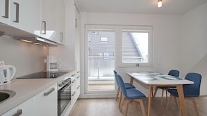Una cocina moderna con armarios blancos, una estufa y una cafetera. Hay una mesa de comedor con sillas azules y vista a un edificio a través de una ventana.
