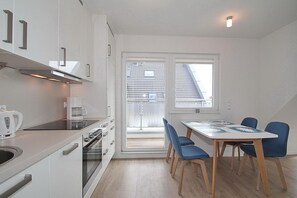 A modern kitchen with white cabinets, a stove, and a coffee maker. There is a dining table with blue chairs and a view of a building through a window.