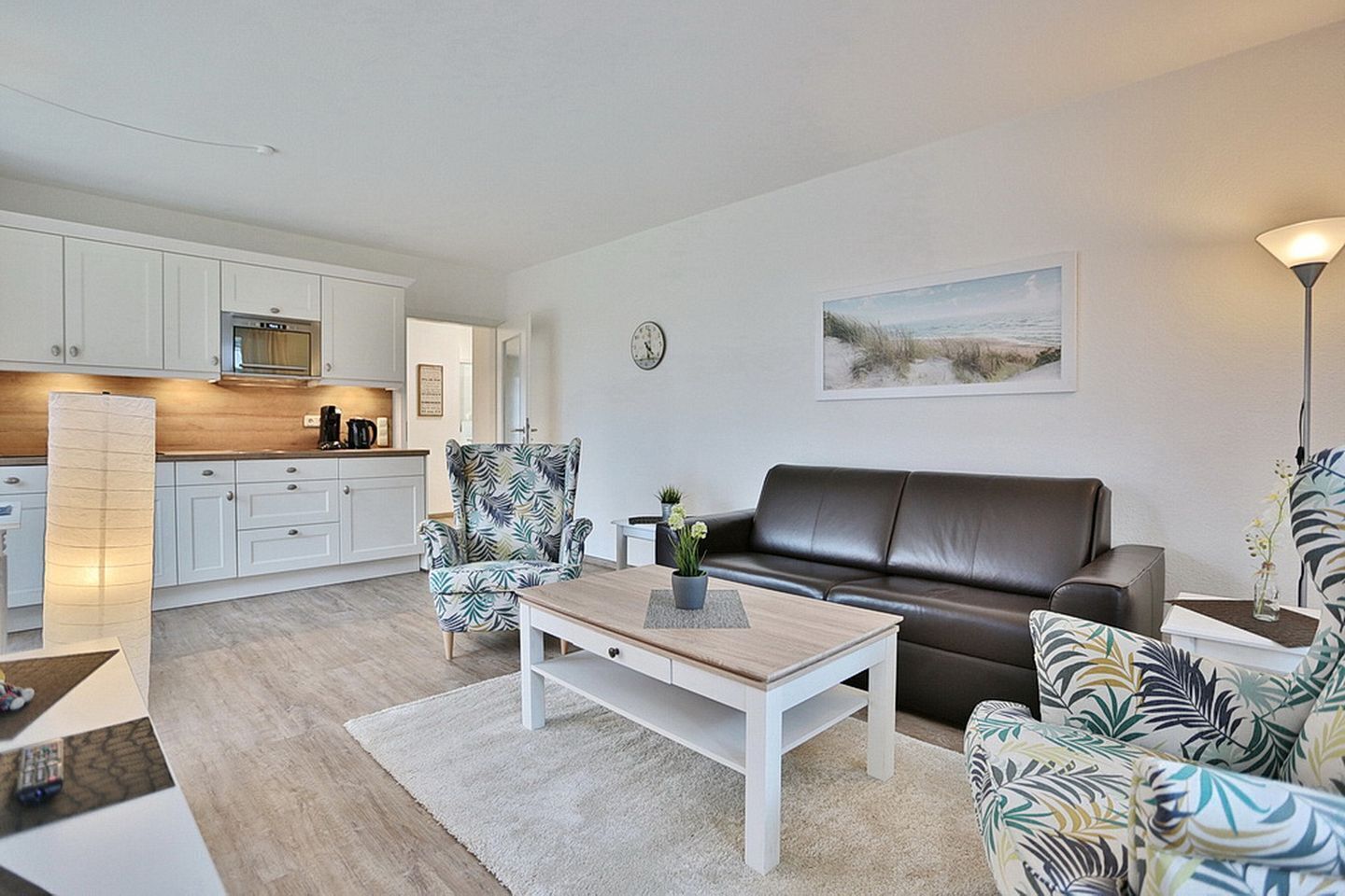 Una sala de estar moderna con un sofá, un sillón y una mesa de centro. Hay una zona de cocina con armarios blancos y un microondas. Un reloj está en la pared, y un cuadro sobre el sofá.