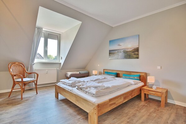 A bedroom with a sloped ceiling, a wooden bed, a chair, a bedside table with a lamp, and a window with curtains.