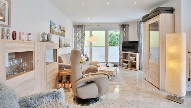 Ein modernes Wohnzimmer mit Sofa, Sessel und TV-Möbel. Eine Glastruhe beherbergt Weingläser und ein dekoratives Objekt.
