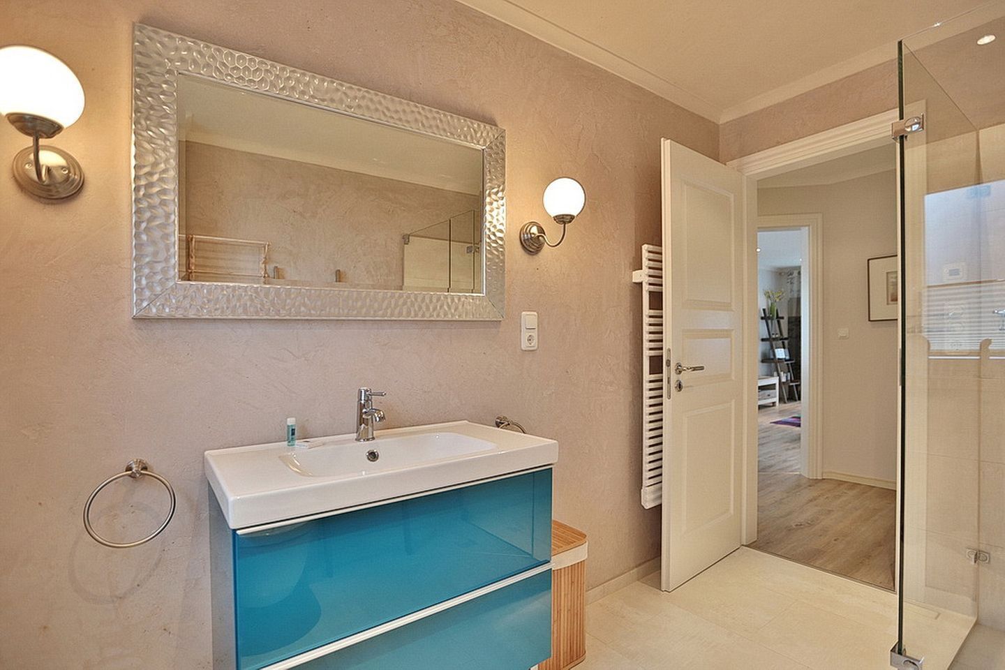 A bathroom with a white sink, blue vanity, and a large mirror. There are wall sconces and a glass shower door.