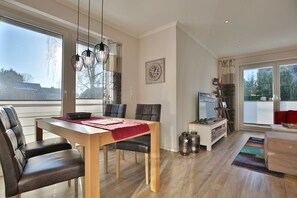 Una sala de estar moderna con una mesa de comedor de madera, sillas de cuero, un televisor y ventanas amplias que ofrecen vistas al exterior.