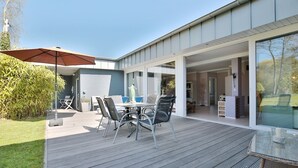 Ein modernes Haus mit einer Terrassenfläche, komplett mit Tisch, Stühlen und einem großen Sonnenschirm, sowie einer Glasdoor, die zu einem Innenbereich führt.