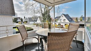 Un balcón con sillas de mimbre y una mesa, con vista a una zona residencial con casas y árboles.