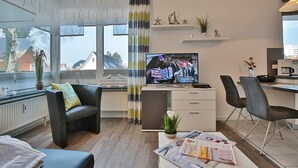 Una sala moderna con televisor, un comedor con sillas, una zona de cocina y vistas al exterior a través de grandes ventanales.