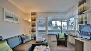 Una sala de estar moderna con sofá, sillón, mesa de centro y televisor. Hay una ventana con cortinas, un radiador y vistas al exterior.