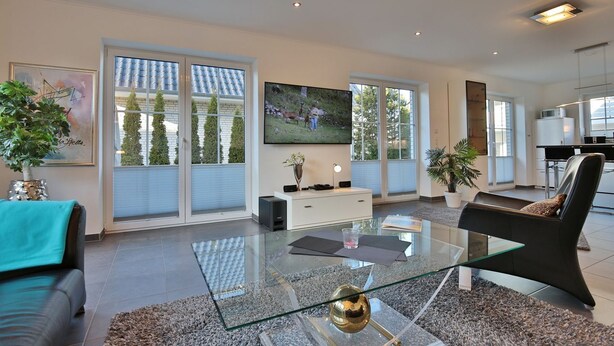 A modern living room with a glass coffee table, a sofa, a TV, and a kitchen in the background.