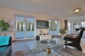 A modern living room with a glass coffee table, a sofa, a TV, and a kitchen in the background. - Villa Mare Apartment 1 - Villa Mare (Timmendorfer Strand)