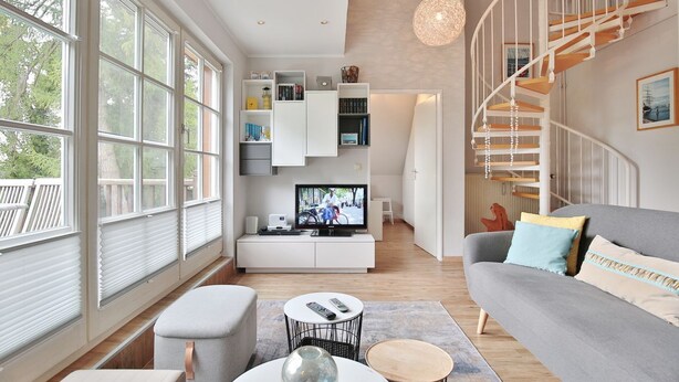 A modern living room with a large window, a grey sofa, a white entertainment unit, and a spiral staircase. - Am Rosenhain 6 Apartment 5 - Am Rosenhain 6 (Timmendorfer Strand)