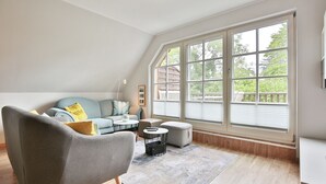 A modern living room with a grey armchair, a light blue sofa, and a coffee table. There is a large window with blinds, and a wooden deck is visible outside. - Am Rosenhain 6 Apartment 5 - Am Rosenhain 6 (Timmendorfer Strand)