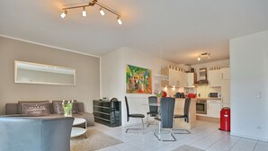 A modern living room with a sofa, a dining table with chairs, a kitchen area with cabinets and appliances, and a painting on the wall. - Strandstrasse 150 Apartment 02 - House Strandstrasse (Timmendorfer Strand)