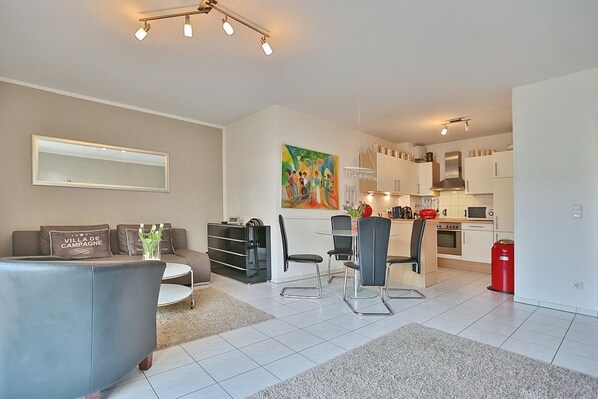 A modern living room with a sofa, a dining table with chairs, a kitchen area with cabinets and appliances, and a painting on the wall. - Strandstrasse 150 Apartment 02 - House Strandstrasse (Timmendorfer Strand)