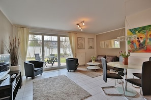 A modern living room with a glass dining table, black chairs, a sofa, and a coffee table. There is a large window with curtains, a radiator, and a painting on the wall. - Strandstrasse 150 Apartment 02 - House Strandstrasse (Timmendorfer Strand)