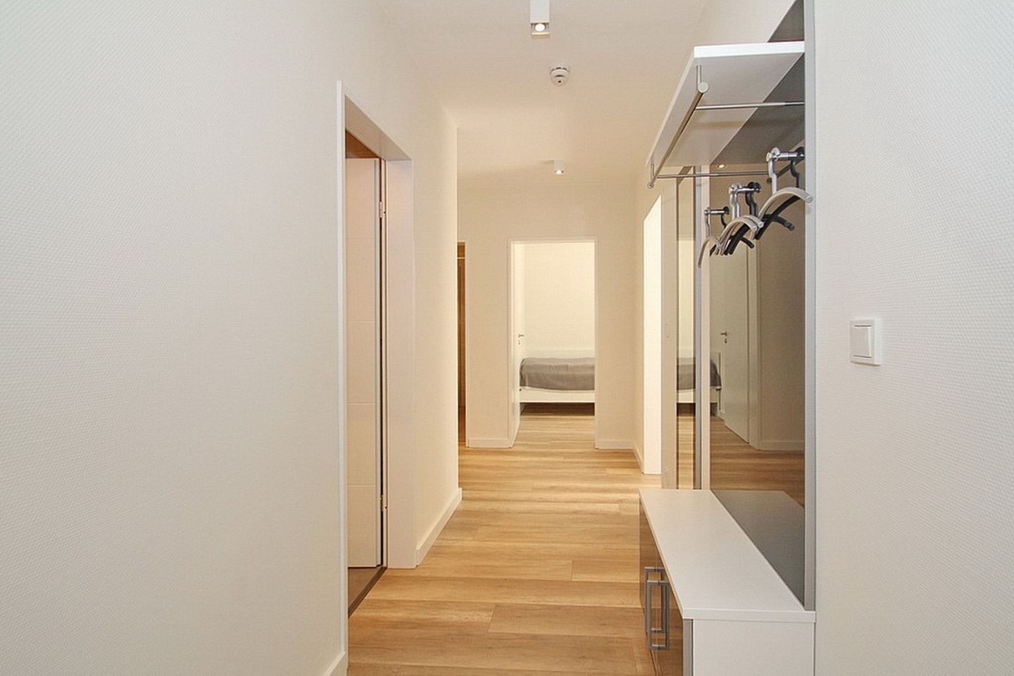 A narrow hallway with wooden flooring leading to a bedroom with a bed and a bathroom with a shower and a mirror.
